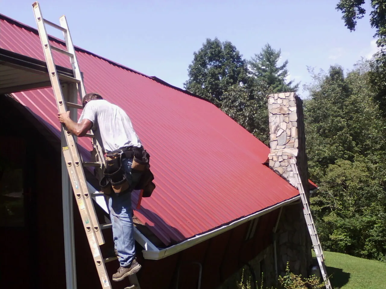 Roofer on a ladder beside a red metal roof and stone chimney — Soffit & Fascia Repair by Roofing Brothers in Beverly Shores